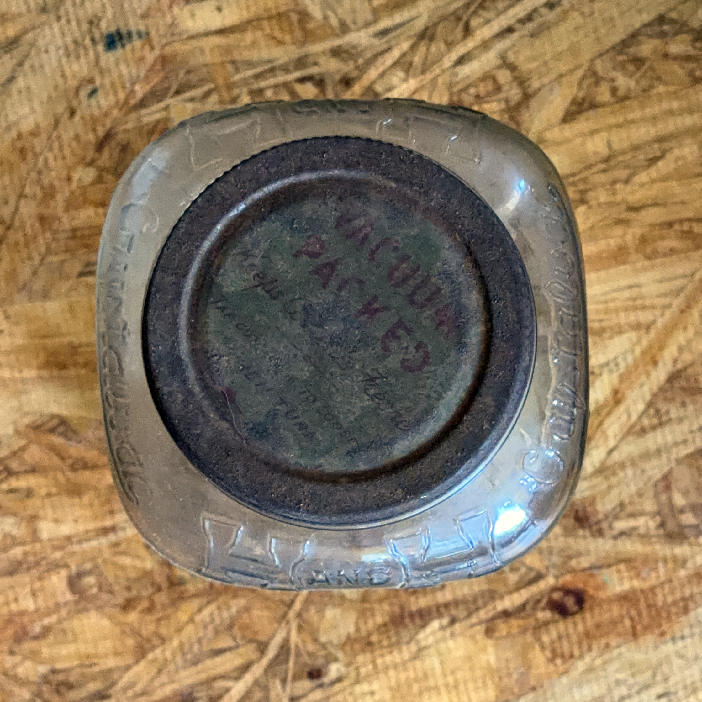 Top-down view of the jar's brown oxidized metal screw lid printed VACUUM PACKED in red, set in the embossed square glass rim