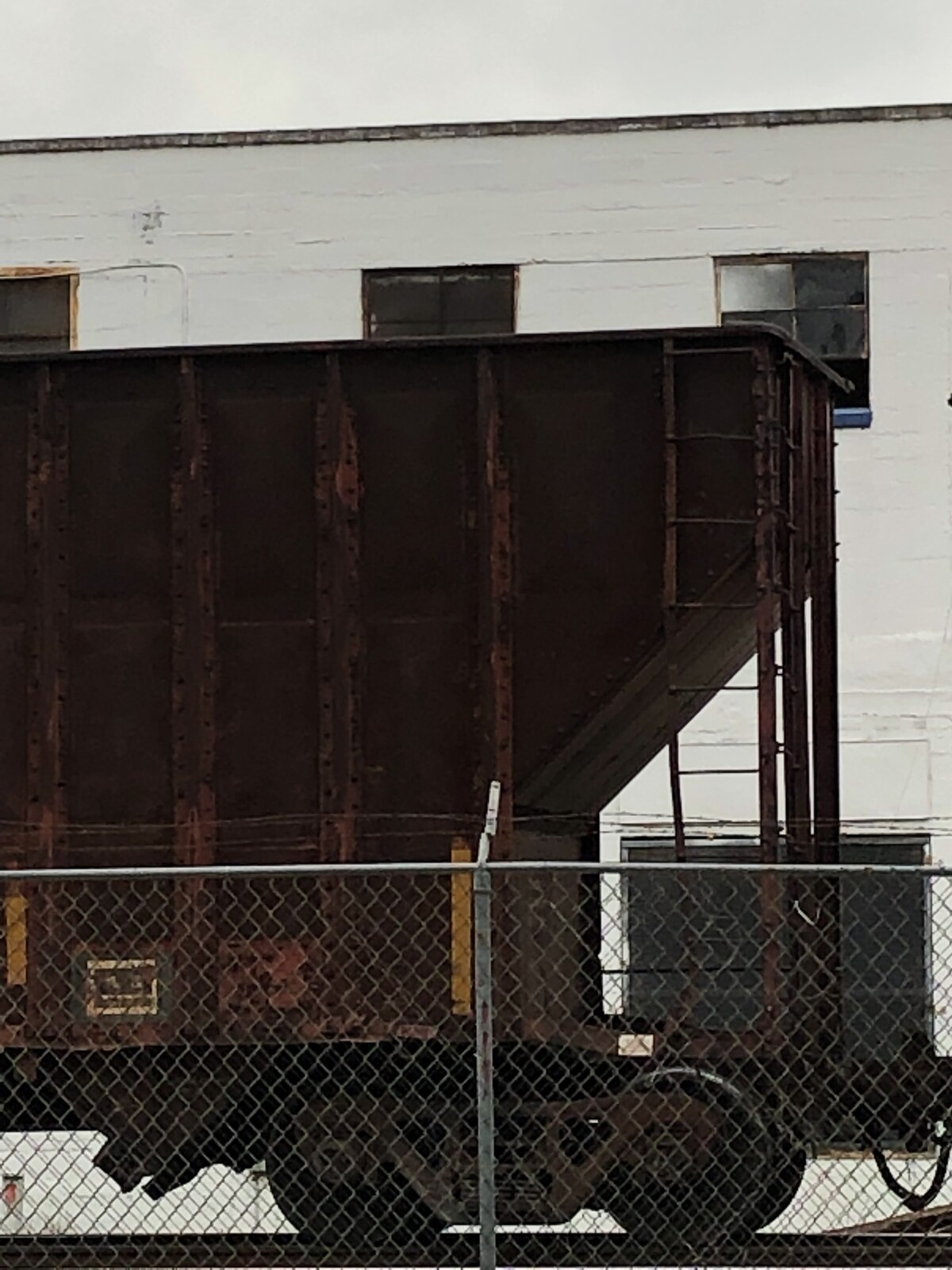 Covered hopper car parked on the rail siding directly behind the 1932 Hoffmann-Hayman Coffee Co. factory wall in San Antonio, photographed through the chain-link fence on an overcast April 2019 afternoon.