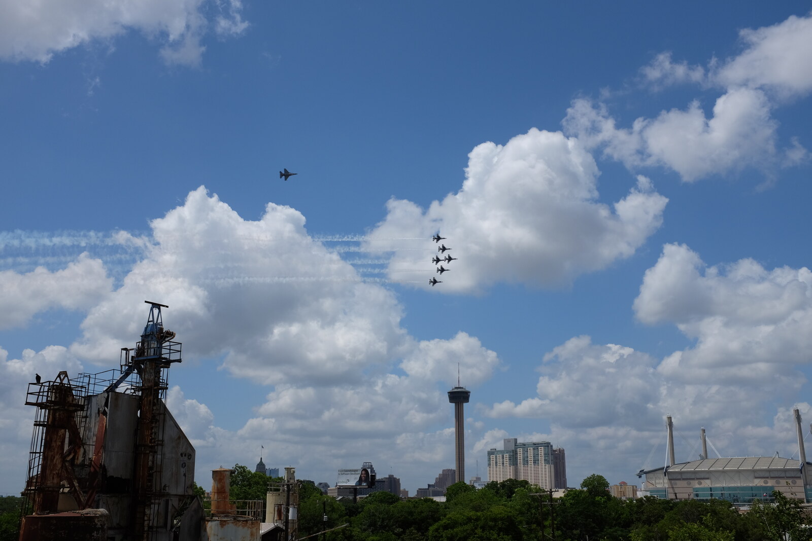 View north from the roof of the 1932 Hoffmann-Hayman Coffee Co. factory on 13 May 2020, with a vulture perched on the railing of the 1008 Hoefgen concrete structure (Alamo Pavers) in the lower-left foreground, the downtown San Antonio skyline and the Tower of the Americas at center, and the USAF Thunderbirds F-16 demonstration team in tight delta formation streaming contrails above the Tower with the solo ship running independently to the upper-left — the team's 'America Strong' COVID-era salute flyover.