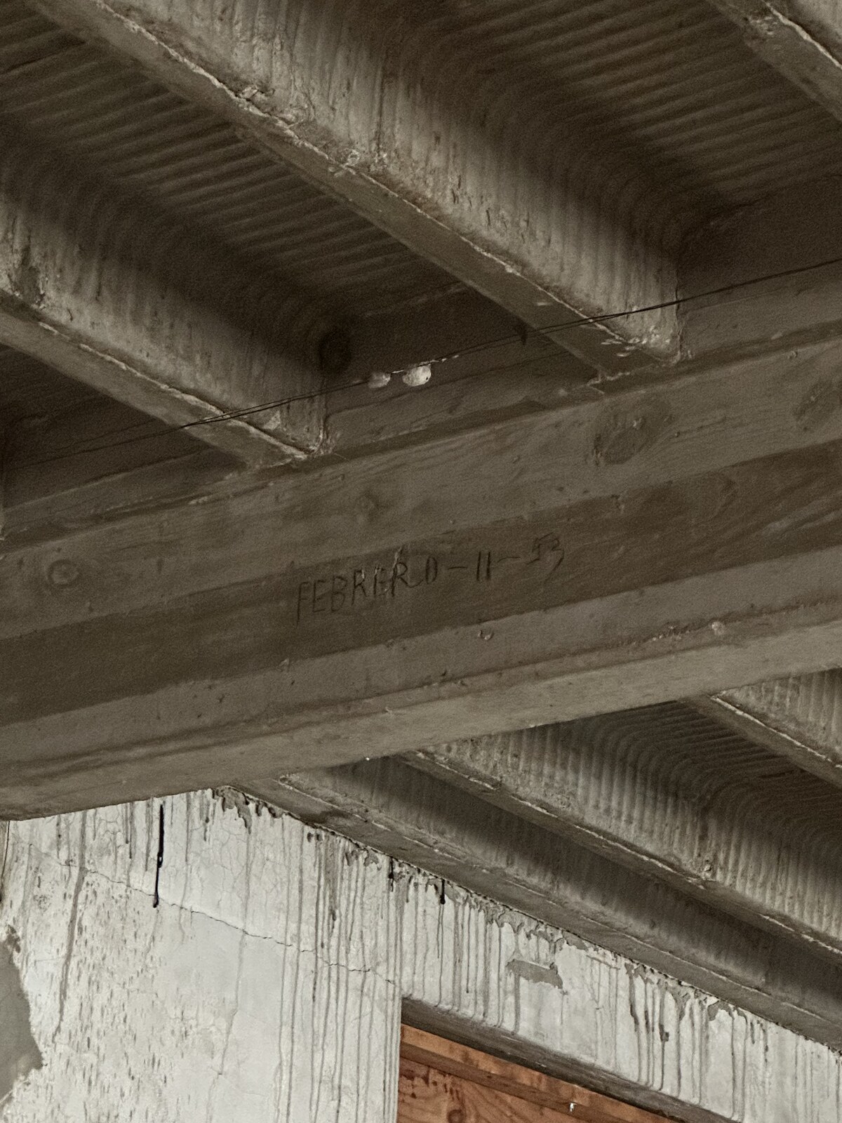 Upward view on 1 February 2023 showing the junction between the pan-formed reinforced-concrete joist ceiling of the second-floor expansion room above the office and one of the original 1932 exterior masonry faces of the plant below — board-formed concrete joists with knot-hole imprints and ribbed corrugated-pan soffit bays, a cursive graphite-pencil inscription on one central joist reading "FEBRERO - 11 - 53" (February 11, 1953), and long vertical whitewash drip-runs cascading down the former outside wall below the ceiling, with a wooden jamb visible at lower-right in a cut opening.