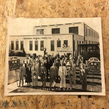 The second press print in the lot: "In Step with N.R.A." — a group of roughly thirty Hoffmann-Hayman employees posed on the factory's front steps, with NRA eagle emblems and a "We are Satisfied — Hoffmann-Hayman Coffee Co." placard on the facade, flanked by two circular "BUY NOW — H-H Brand Quality Products" display signs on staffs. An H and H Coffee delivery truck is parked on the right. Caption "IN STEP WITH N.R.A." runs along the foreground.