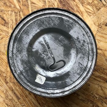 Top-down view of the plain grey-painted lid, with the original unused keywind tool soldered at center — a flat blade with an eye at the base to feed the lid strip through — and a small hand-written price sticker toward the bottom edge.